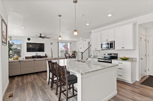 kitchen with appliances with stainless steel finishes, white cabinetry, hanging light fixtures, light stone counters, and a center island with sink