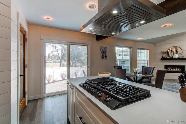 kitchen with island exhaust hood, black gas cooktop, a warm lit fireplace, wood finished floors, and white cabinetry