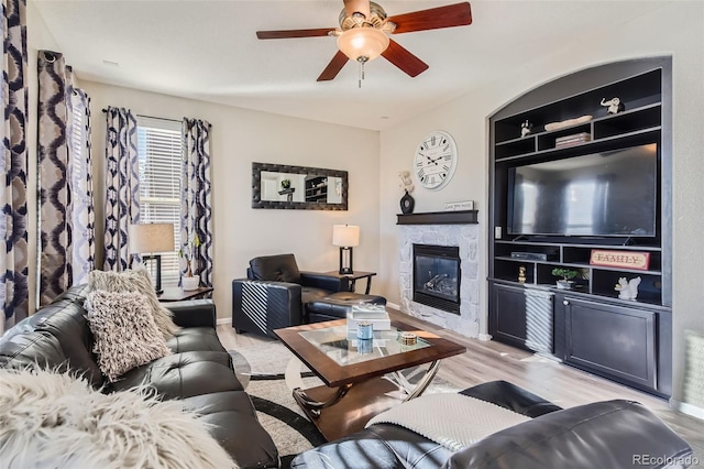 living area featuring light wood-style flooring, a ceiling fan, baseboards, built in features, and a glass covered fireplace