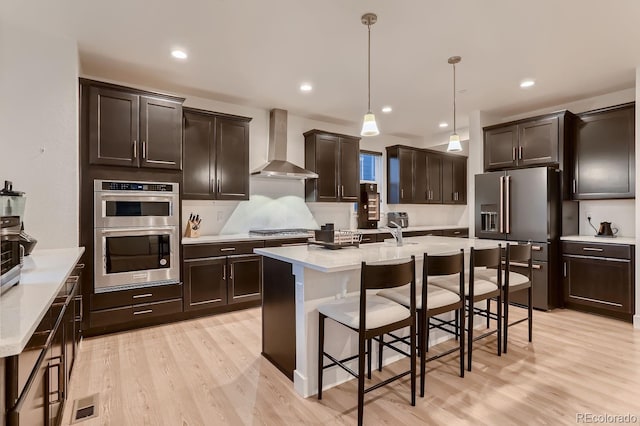 kitchen featuring pendant lighting, light wood-type flooring, wall chimney exhaust hood, appliances with stainless steel finishes, and dark brown cabinetry