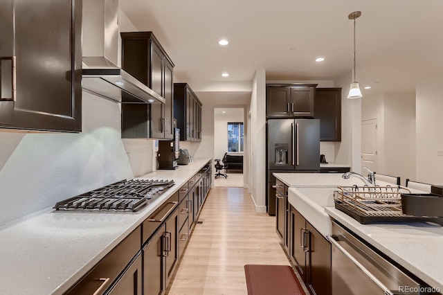 kitchen featuring pendant lighting, dark brown cabinets, wall chimney range hood, appliances with stainless steel finishes, and light wood-type flooring