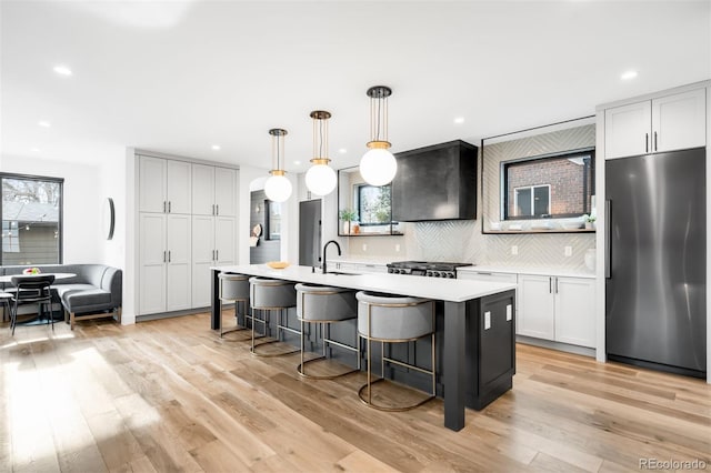 kitchen featuring light countertops, a center island with sink, backsplash, stainless steel refrigerator, and recessed lighting