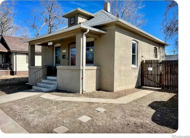 view of front of house with stucco siding, fence, roof with shingles, covered porch, and a chimney