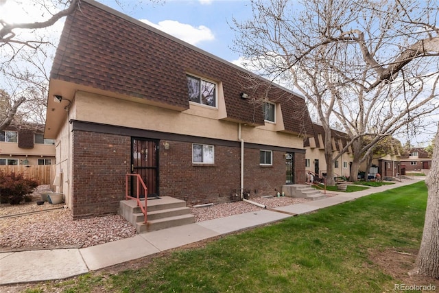 rear view of house with brick siding, mansard roof, a yard, a shingled roof, and stucco siding