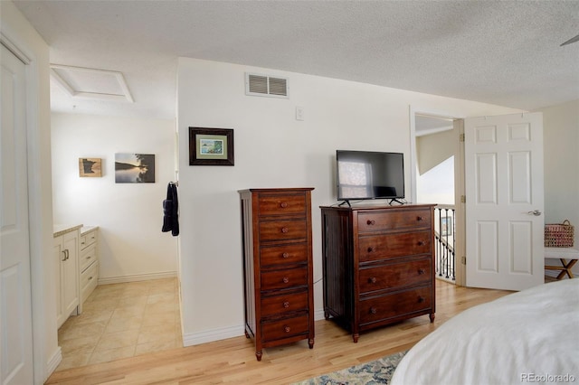 bedroom with a textured ceiling, light wood-style flooring, visible vents, baseboards, and attic access