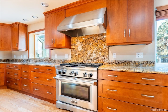 kitchen featuring wall chimney exhaust hood, light stone counters, light wood-type flooring, high end stainless steel range oven, and decorative backsplash