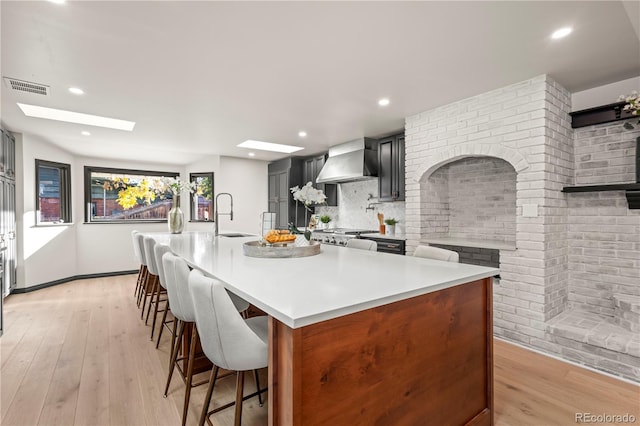 kitchen featuring a kitchen island with sink, wall chimney exhaust hood, stainless steel stove, light wood-type flooring, and tasteful backsplash