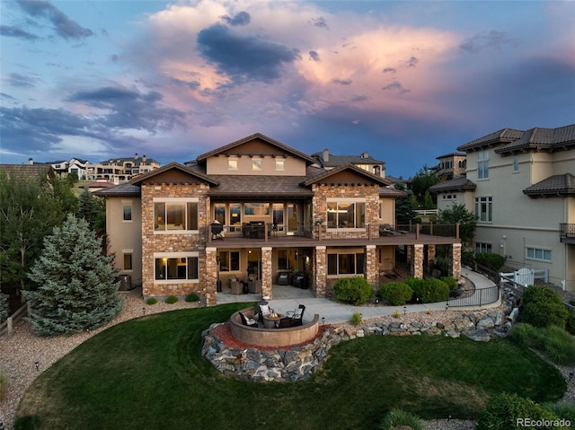 back of house at dusk featuring an outdoor fire pit, stone siding, a lawn, stucco siding, and a patio area