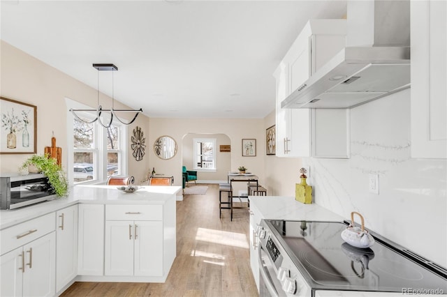 kitchen with white cabinets, wall chimney exhaust hood, appliances with stainless steel finishes, decorative light fixtures, and light hardwood / wood-style floors