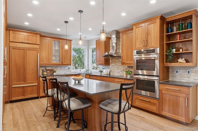 kitchen with recessed lighting, light wood finished floors, stainless steel double oven, wall chimney exhaust hood, and a kitchen bar