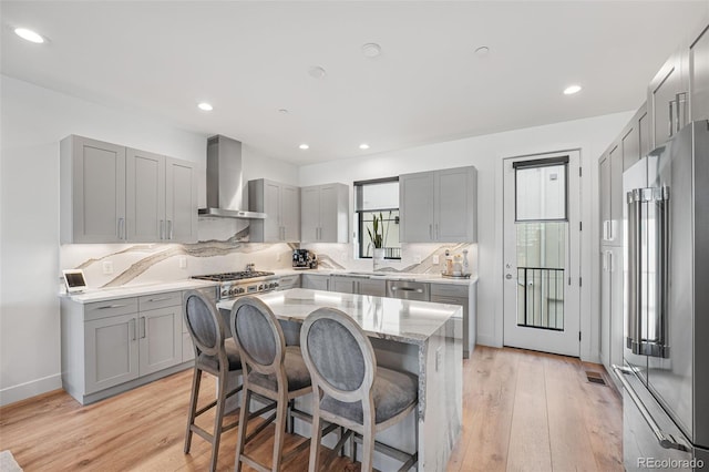 kitchen with high end refrigerator, wall chimney range hood, light wood-type flooring, gray cabinets, and tasteful backsplash