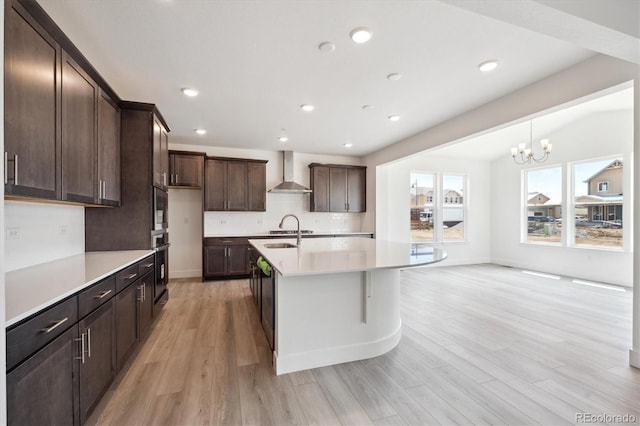 1376 Lumber Ridge Cir N, Erie, CO 80516 - kitchen featuring wall chimney range hood, light countertops, light wood-style flooring, dark brown cabinets, and a kitchen island with sink