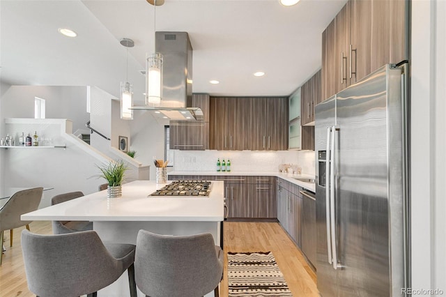 kitchen featuring hanging light fixtures, stainless steel appliances, island exhaust hood, decorative backsplash, and light wood-type flooring