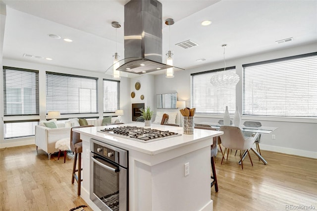 kitchen featuring appliances with stainless steel finishes, island range hood, decorative light fixtures, a kitchen bar, and light hardwood / wood-style flooring