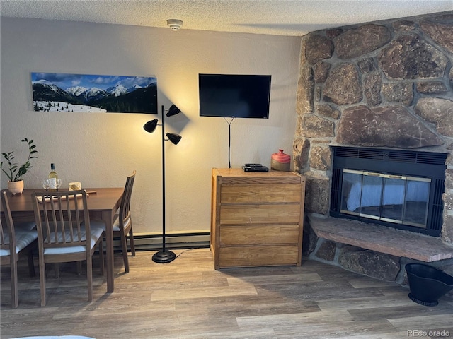 dining space featuring a textured wall, a baseboard radiator, wood finished floors, a textured ceiling, and a stone fireplace