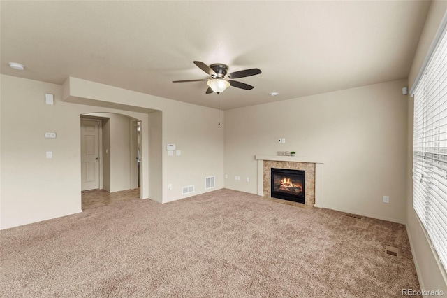 unfurnished living room with a ceiling fan, light colored carpet, arched walkways, and a tiled fireplace