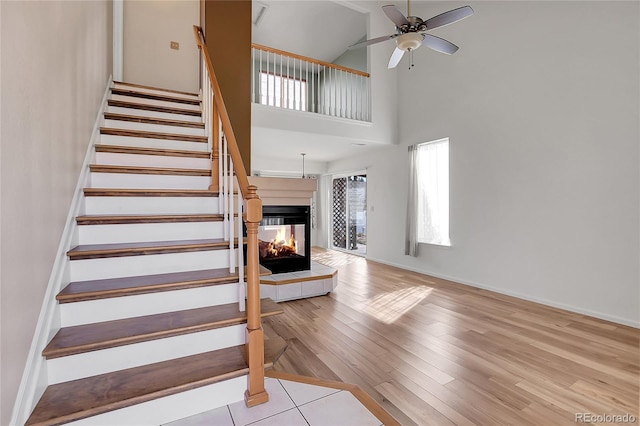 stairway with a ceiling fan, wood finished floors, a multi sided fireplace, a high ceiling, and baseboards