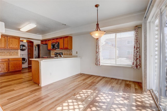 kitchen with brown cabinetry, light wood-type flooring, stainless steel appliances, stacked washer / drying machine, and tasteful backsplash