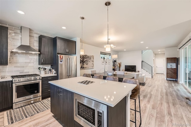 kitchen featuring wall chimney range hood, premium appliances, pendant lighting, a kitchen island, and light wood-type flooring