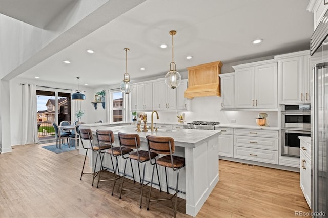 kitchen with white cabinets, a center island with sink, appliances with stainless steel finishes, light hardwood / wood-style flooring, and decorative light fixtures