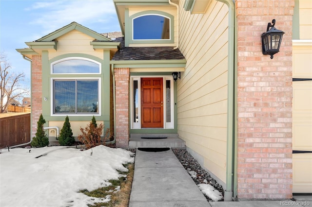 snow covered property entrance with brick siding, roof with shingles, and fence