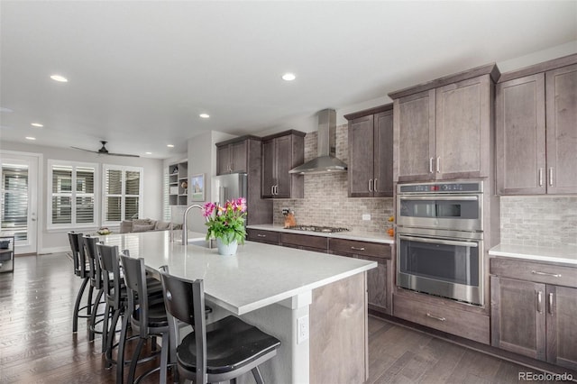 15843 E Broncos Pl, Englewood, CO 80112 - kitchen featuring wall chimney exhaust hood, stainless steel appliances, dark wood-type flooring, a kitchen bar, and a kitchen island with sink