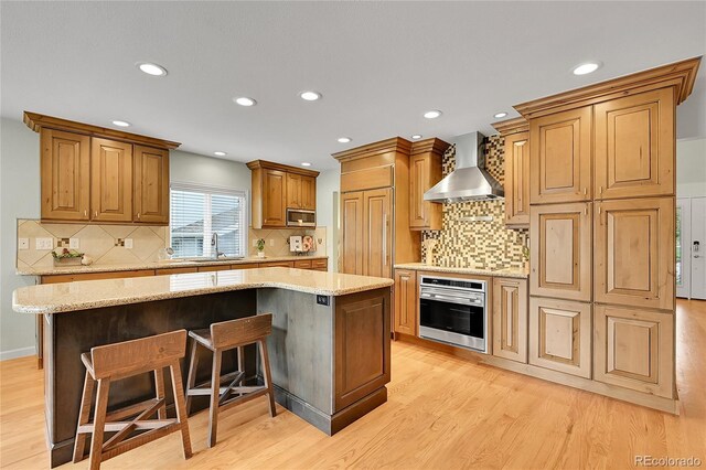 kitchen featuring appliances with stainless steel finishes, wall chimney range hood, a sink, light wood-style flooring, and a kitchen island