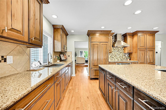 kitchen featuring wall chimney range hood, a sink, light stone countertops, light wood-type flooring, and backsplash