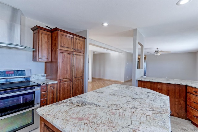 kitchen featuring ceiling fan, wall chimney range hood, stainless steel electric stove, a kitchen island, and light wood-type flooring