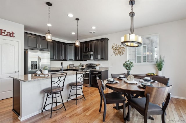 kitchen with appliances with stainless steel finishes, decorative light fixtures, light stone counters, light hardwood / wood-style flooring, and a center island with sink