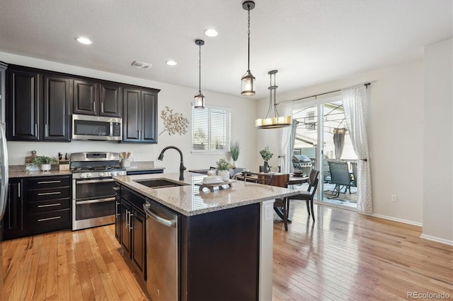 kitchen featuring decorative light fixtures, stainless steel appliances, an island with sink, sink, and light wood-type flooring