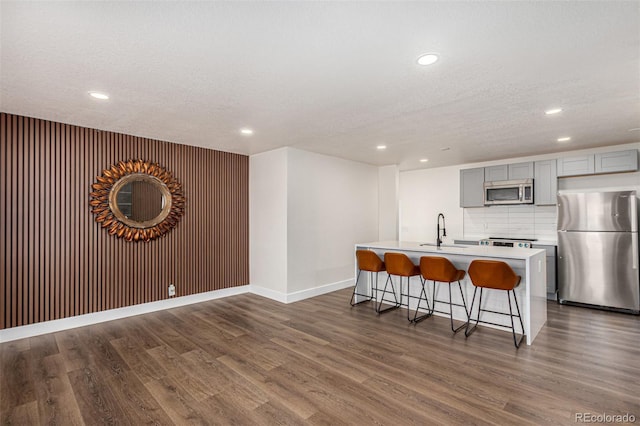 kitchen featuring gray cabinetry, a center island with sink, a sink, dark wood-style floors, and appliances with stainless steel finishes