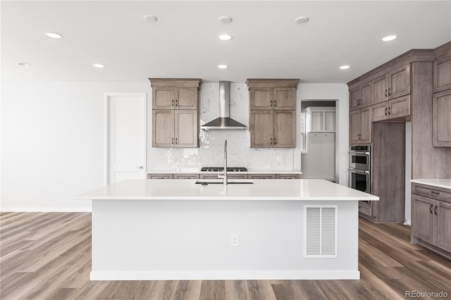 8858 Moonshimmer Trl, Littleton, CO 80125 - kitchen featuring wall chimney range hood, stainless steel double oven, an island with sink, backsplash, and wood finished floors