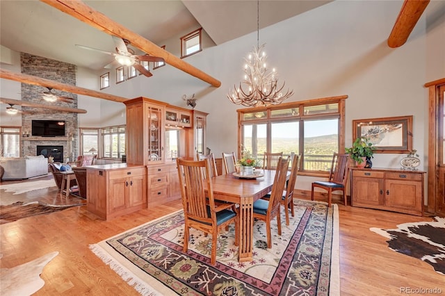 dining space featuring ceiling fan with notable chandelier, a fireplace, a high ceiling, light hardwood / wood-style floors, and beam ceiling