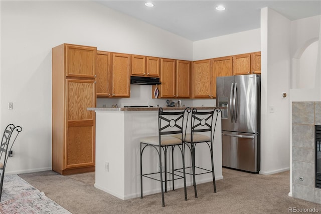 kitchen featuring stainless steel refrigerator with ice dispenser, light carpet, under cabinet range hood, a kitchen bar, and baseboards