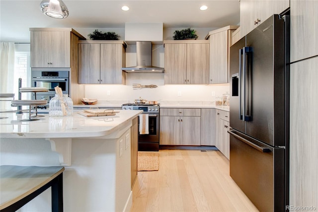 kitchen with wall chimney exhaust hood, light stone counters, light hardwood / wood-style floors, light brown cabinets, and stainless steel appliances