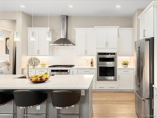 kitchen featuring white cabinets, wall chimney exhaust hood, appliances with stainless steel finishes, light wood finished floors, and decorative backsplash