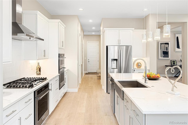kitchen featuring wall chimney range hood, an island with sink, backsplash, stainless steel appliances, and light wood finished floors