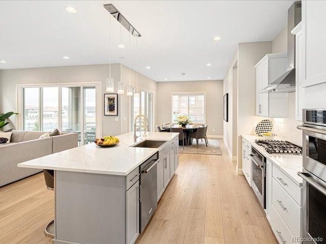 kitchen featuring light wood-type flooring, wall chimney range hood, a kitchen island with sink, stainless steel appliances, and a sink