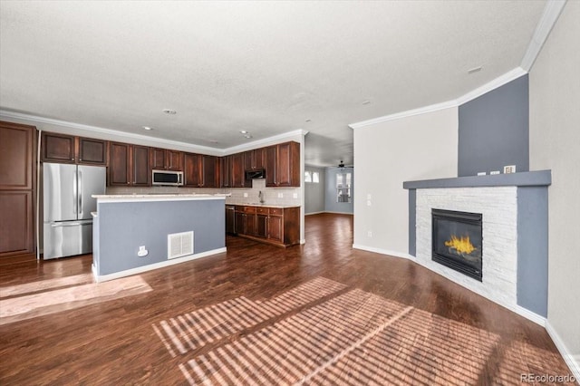 kitchen featuring crown molding, dark wood-type flooring, stainless steel appliances, a kitchen island, and a stone fireplace