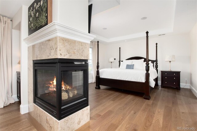 bedroom with a tray ceiling, light wood-type flooring, and a multi sided fireplace