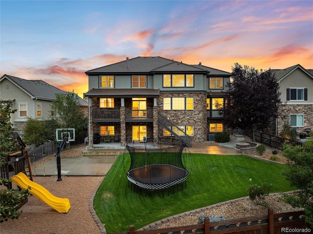 back house at dusk featuring a trampoline, a playground, a patio, and a lawn