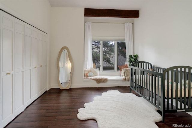 bedroom featuring a closet, beamed ceiling, baseboards, dark wood-style floors, and a crib