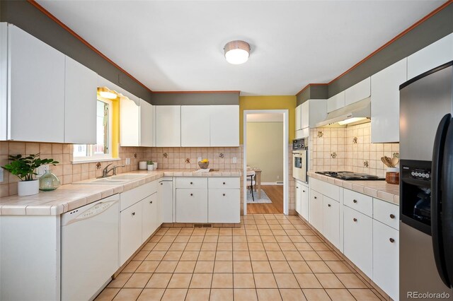 kitchen featuring white dishwasher, a sink, tile countertops, stainless steel fridge with ice dispenser, and under cabinet range hood