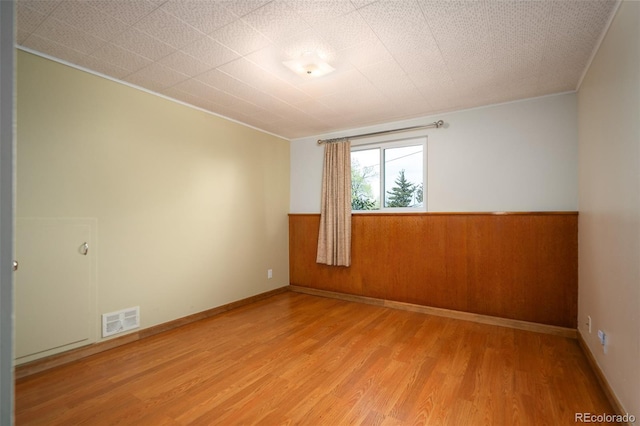 empty room with crown molding, a wainscoted wall, light wood finished floors, wood walls, and visible vents