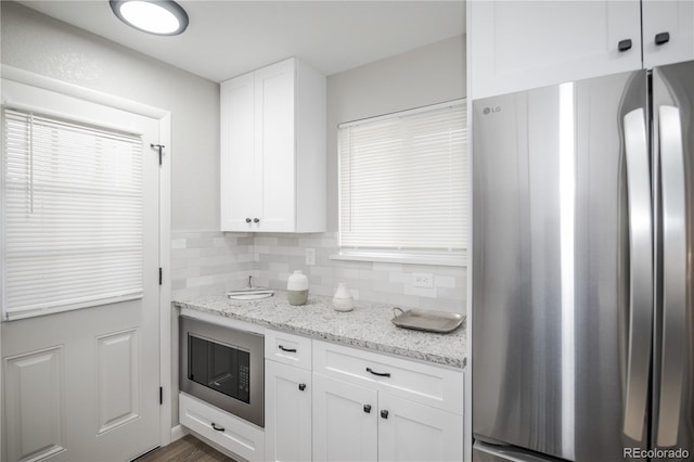 kitchen with decorative backsplash, light stone counters, white cabinetry, black microwave, and stainless steel refrigerator