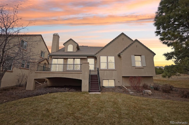 back of house at dusk with stairs, a lawn, a wooden deck, stucco siding, and a chimney