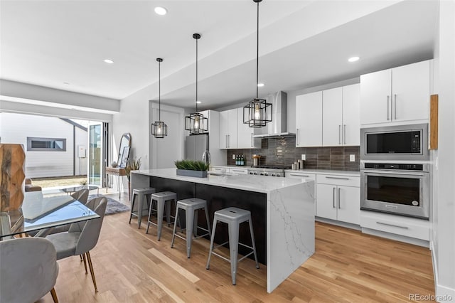 kitchen featuring a kitchen island with sink, wall chimney range hood, stainless steel appliances, and white cabinets