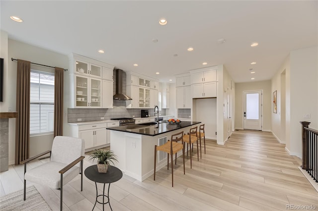 kitchen with high end stainless steel range, white cabinets, light wood-type flooring, wall chimney range hood, and a center island with sink