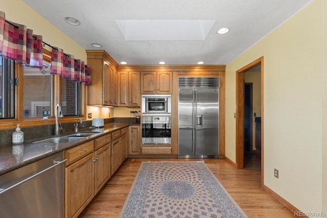 kitchen with sink, appliances with stainless steel finishes, a skylight, dark stone counters, and light wood-type flooring